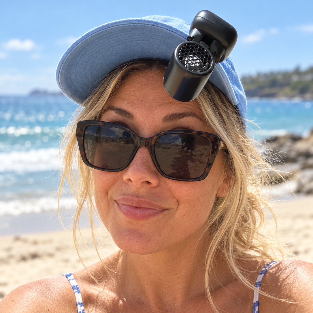 A woman on a sunny beach wearing a light blue baseball cap with a black handheld fan attached to the brim, dark sunglasses, and a blue-and-white patterned bikini top.
