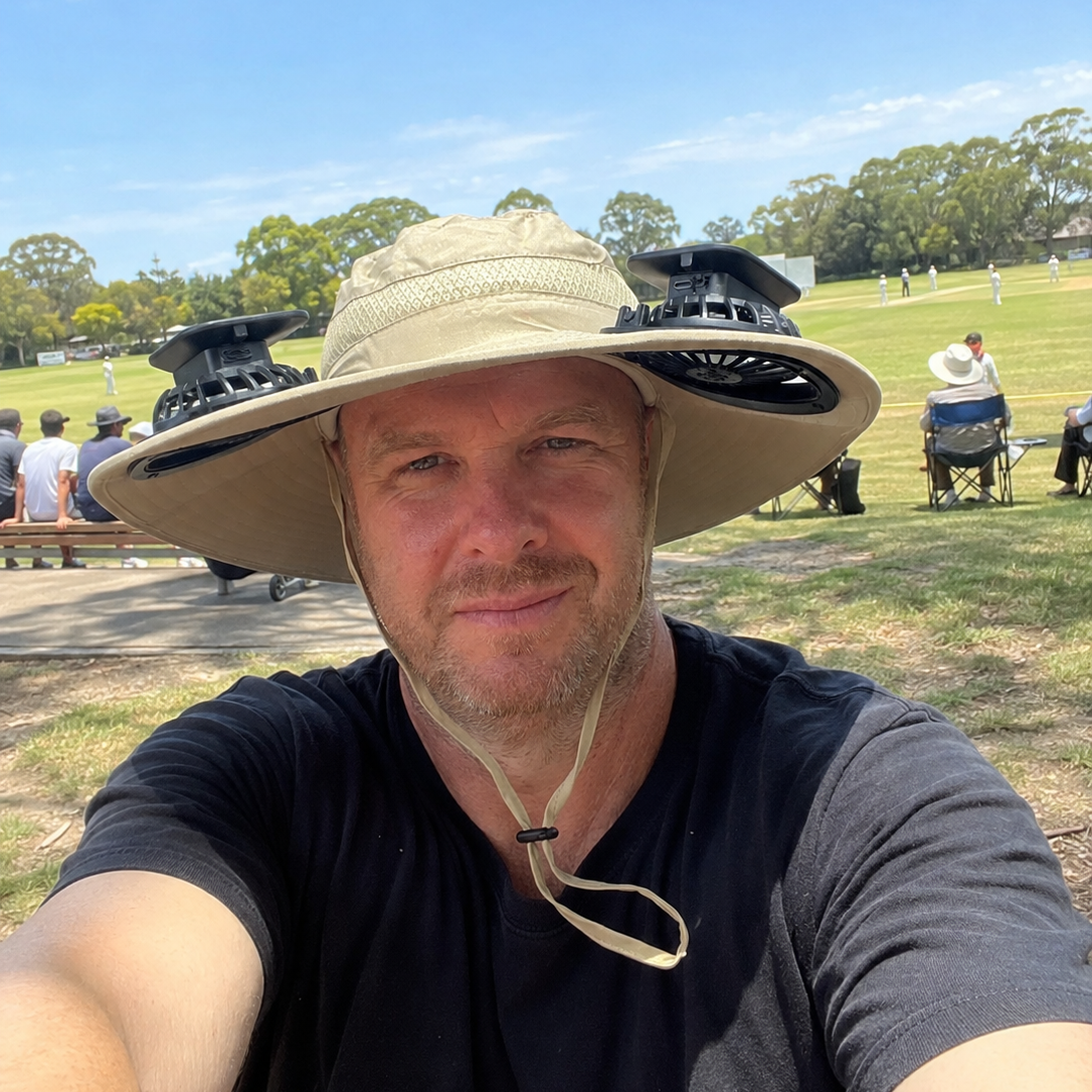 A man taking a selfie outdoors on a sunny day, wearing a beige wide-brim sun hat with black cooling fans attached to the brim and crown, along with a black t-shirt. The background shows a grassy park with trees and other people under a blue sky.