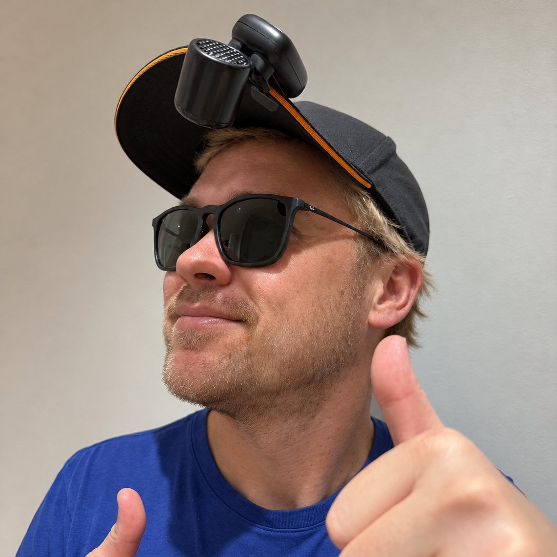 A man wearing a black and orange baseball cap with a mounted camera on the brim, black sunglasses, and a blue t-shirt, giving a thumbs-up against a plain gray wall.