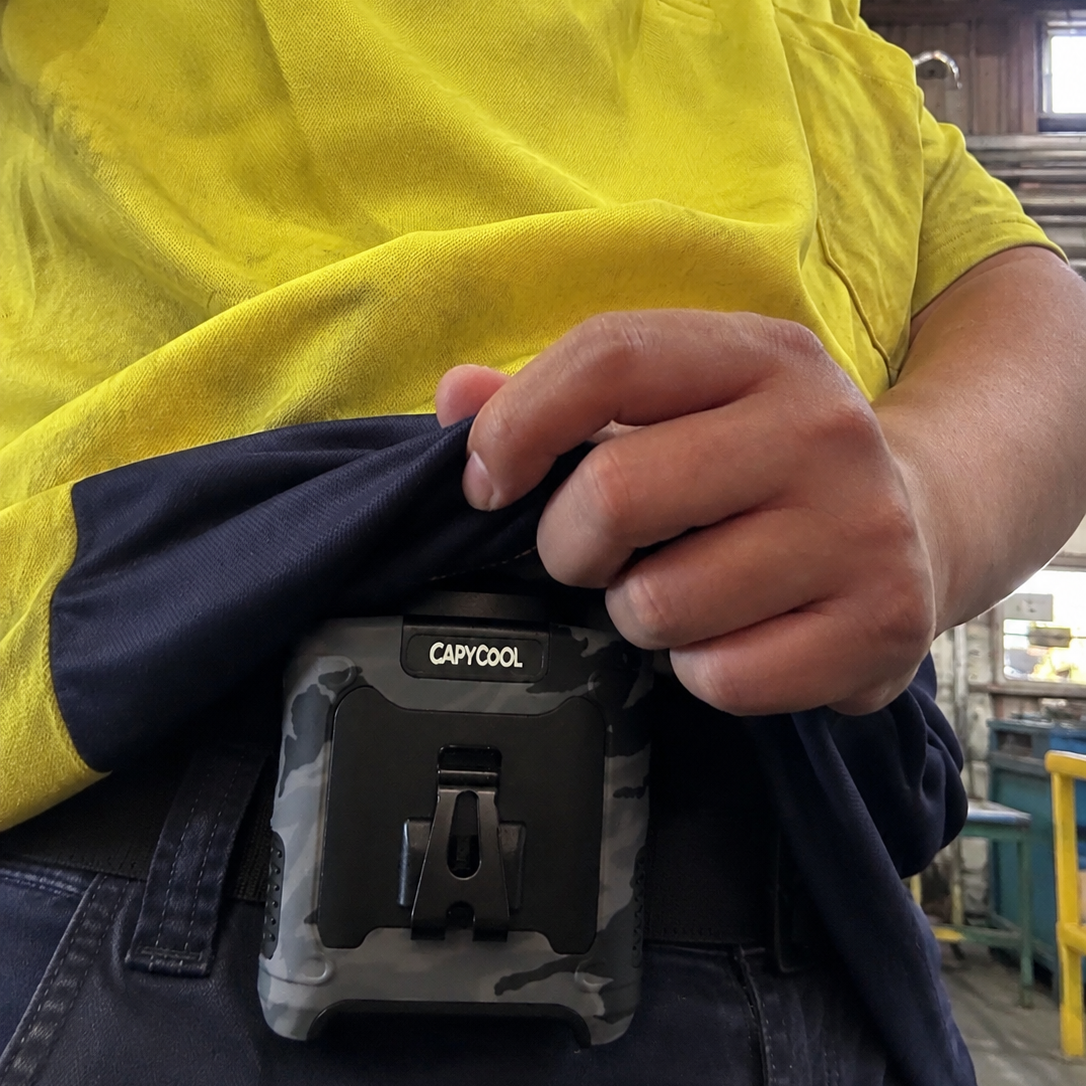 A close-up of a person wearing a yellow and navy work shirt, showing a CAPCOOL rugged fan mounted on their belt buckle.
